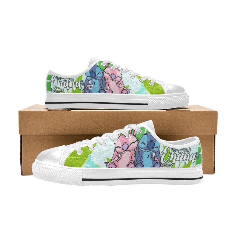 Puede incluir: Zapatillas blancas de lona con una imagen de dibujos animados de Stitch y Angel de Lilo & Stitch. Las zapatillas tienen un fondo verde y azul con la palabra "Ohana" impresa en el lateral.
