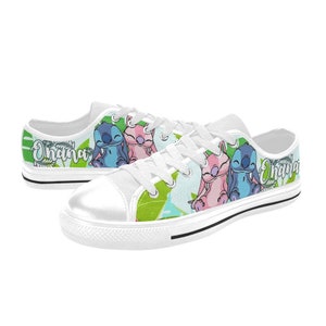 Puede incluir: Zapatillas bajas de lona blancas con un estampado colorido de Stitch y Angel de Lilo & Stitch. El estampado incluye hojas verdes, personajes azules y rosas, y la palabra "Ohana" en ambos zapatos.