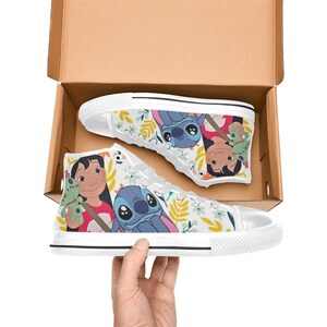 May include: White high-top canvas sneakers featuring a colorful cartoon design. The sneakers showcase characters and floral patterns. The shoes are in a cardboard box, with one shoe being held by a hand.