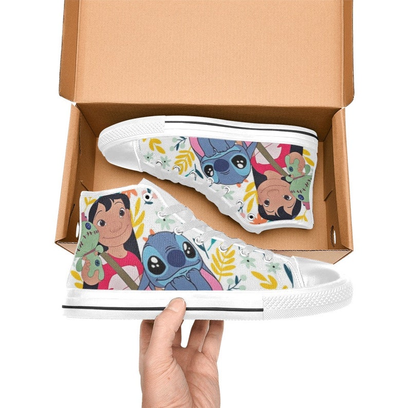 May include: White high-top canvas sneakers featuring a colorful cartoon design. The sneakers showcase characters and floral patterns. The shoes are in a cardboard box, with one shoe being held by a hand.