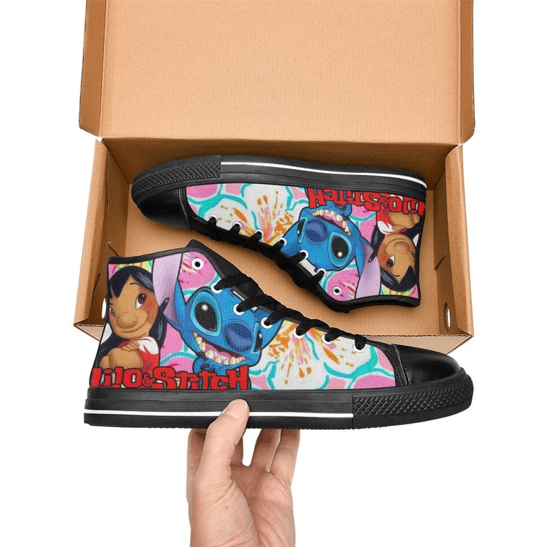 Op de afbeelding: Een paar zwarte high-top canvas schoenen met een kleurrijke print met Stitch en Lilo uit de Disney-film Lilo & Stitch. De print bevat roze bloemen en de tekst "Lilo & Stitch".
