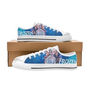 Puede incluir: Zapatillas bajas de lona blancas con un dise&ntilde;o azul y blanco con personajes de la pel&iacute;cula Frozen. Las zapatillas tienen cordones negros y una suela de goma negra. El texto "Frozen" est&aacute; impreso en el lateral de las zapatillas.