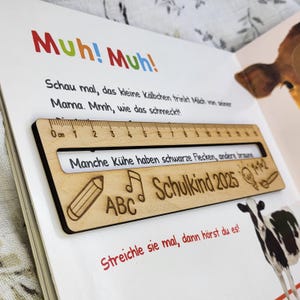 May include: A wooden ruler with a 12 cm scale, resting on a children's book page. The ruler has etched images of a pencil, musical note, and the text "Schulkind 2025". The book's text includes "Muh! Muh!" and a cow illustration.