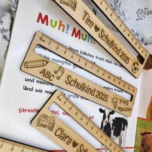 May include: Wooden rulers with engraved text and designs, including names, hearts, and the text "Schulkind 2025." The rulers have a 15 cm scale and are placed on a book with colorful text and illustrations.