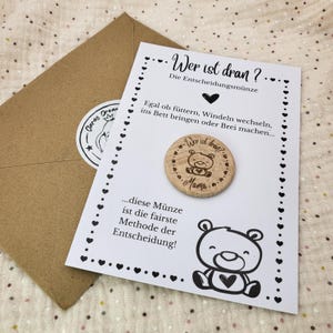 Può includere: Una moneta decisionale in legno con un'illustrazione di orso e la parola "Mama" è su una carta bianca con testo e una grafica di orso. Una busta con un adesivo è sullo sfondo. Il testo sulla carta recita "Wer ist dran?" e "...diese Münze ist die fairste Methode der Entscheidung!"