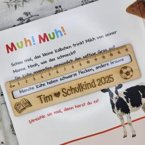 May include: A wooden ruler with a 15-centimeter scale, featuring a soccer ball and book icon, rests on a children's book page. The ruler is engraved with the text "Tim Schulkind 2025". The book has text in German and a cow illustration.