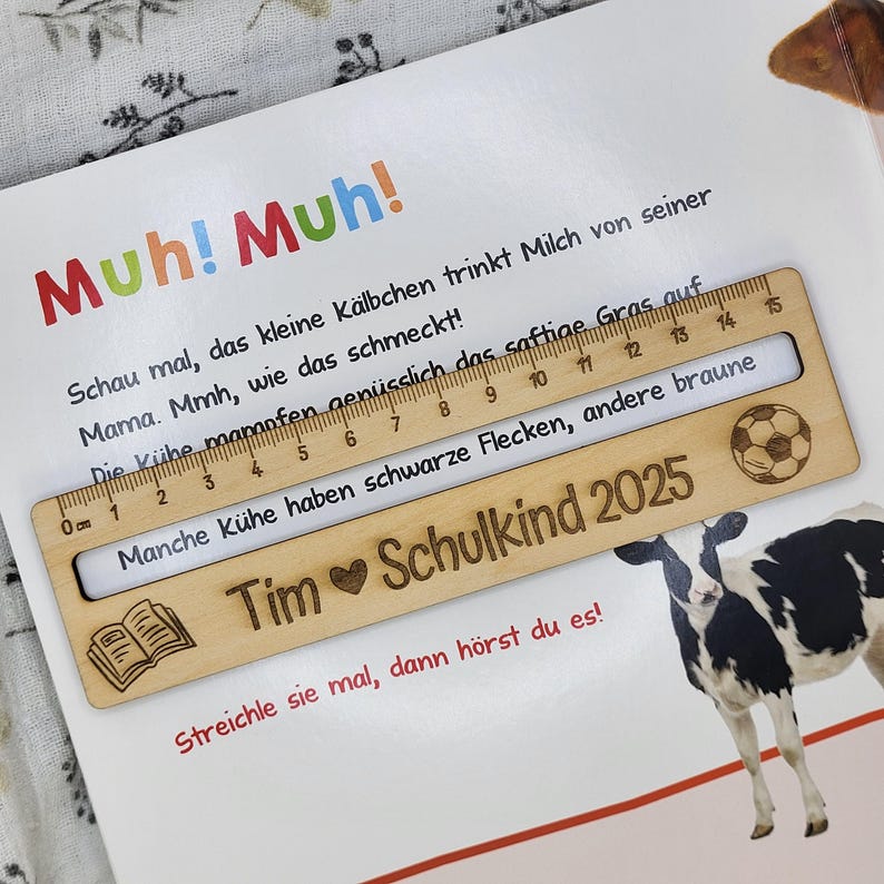 May include: A wooden ruler with a 15-centimeter scale, featuring a soccer ball and book icon, rests on a children's book page. The ruler is engraved with the text "Tim Schulkind 2025". The book has text in German and a cow illustration.