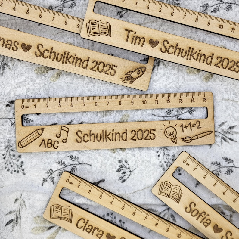 May include: Wooden rulers with engraved names and the text "Schulkind 2025" are displayed. Each ruler has a different name, such as Tim, Clara, and Sofia, along with small engraved icons like a book, pencil, and rocket. The rulers are marked in centimeters.
