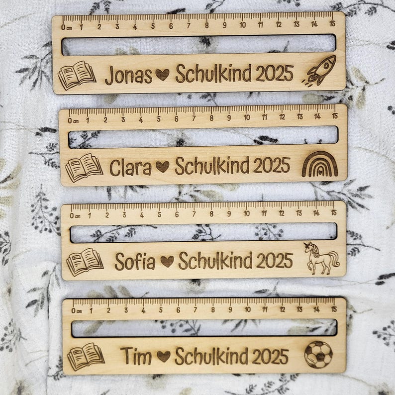 May include: Four wooden rulers, each measuring 15 cm, with engraved text and illustrations. The rulers are personalized with names like "Jonas" and "Clara", along with "Schulkind 2025" and various icons such as a rocket and a unicorn.