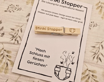 May include: A wooden clothespin with the text "Stinki Stopper" printed on it. The clothespin is designed to look like a diaper with a small, cartoon-style poop emoji on it. The text "Mach Schluss mit fiesen Gerüchen" is printed below the clothespin.