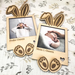 Può includere: Due cornici in legno a tema pasquale. Ogni cornice ha un design a orecchie di coniglio nella parte superiore e zampe di coniglio nella parte inferiore. Una cornice dice "Frohe Ostern" e l'altra "Mein 1. Ostern".