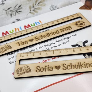 May include: Wooden rulers with engraved text and designs. One ruler has a soccer ball, another has a book icon, and all have measurement markings. The rulers are personalized with names and the text "Schulkind 2025".