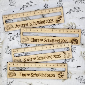 May include: Wooden rulers with engraved names, hearts, and the text "Schulkind 2025". Each ruler has a different icon: a rocket, a rainbow, a unicorn, and a soccer ball. The rulers measure up to 15 cm.