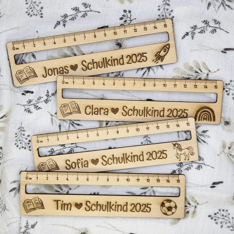 May include: Wooden rulers with engraved names, hearts, and the text "Schulkind 2025". Each ruler has a different icon: a rocket, a rainbow, a unicorn, and a soccer ball. The rulers measure up to 15 cm.