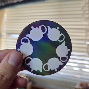 May include: A round sticker with a holographic background featuring a pattern of white teapots arranged in a circle. The sticker is held by a hand, showcasing its design and iridescent effect.