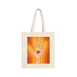 May include: A cream-colored canvas tote bag featuring a central graphic of an open palm with an eye in the center. The hand is decorated with various small symbols on the fingers and wrist, set against a background of wavy orange and brown lines.