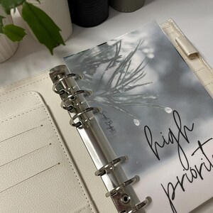 Könnte beinhalten: Ein weißer, ledergebundener Planer mit silbernen Ringen und einem durchsichtigen Plastik-Seitenschutz. Der Seitenschutz zeigt ein Schwarzweißbild eines Kiefernastes mit Schnee und dem Text "high priority".