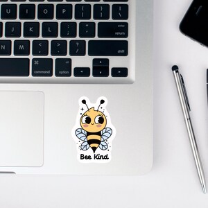 May include: A sticker with a cartoon bee with a yellow body, black stripes, and blue wings. The bee has a smiling face with eyelashes and a pink blush. The text "Bee Kind" is below the bee.