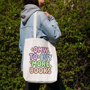 May include: A white tote bag with the text "OMW TO BUY MORE BOOKS" in colourful, outlined letters. The bag has a light blue strap and is being held by a person wearing a light blue denim jacket.