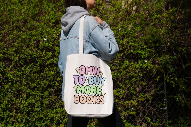May include: A white tote bag with the text "OMW TO BUY MORE BOOKS" in colourful, outlined letters. The bag has a light blue strap and is being held by a person wearing a light blue denim jacket.