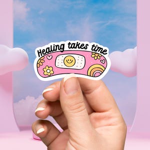 May include: A pink and yellow sticker with a cartoon bandage and the text "Healing takes time".