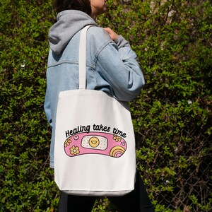 May include: White tote bag with a pink and yellow band-aid design and the text "Healing takes time".