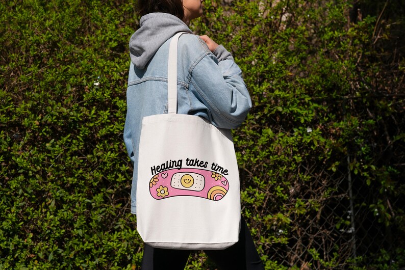 May include: White tote bag with a pink and yellow band-aid design and the text "Healing takes time".
