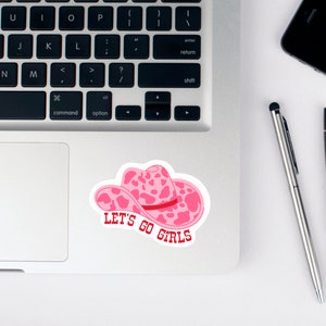 Pode incluir: Um autocolante de chap&eacute;u de cowboy rosa com um design de estampa de vaca e o texto "LET'S GO GIRLS" em vermelho. O autocolante est&aacute; num laptop, com um smartphone preto e canetas por perto.
