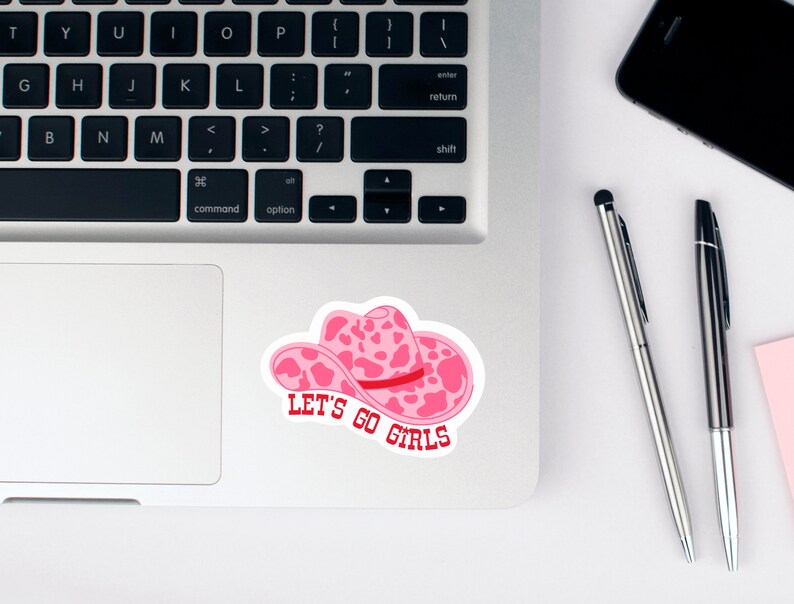 Pode incluir: Um autocolante de chap&eacute;u de cowboy rosa com um design de estampa de vaca e o texto "LET'S GO GIRLS" em vermelho. O autocolante est&aacute; num laptop, com um smartphone preto e canetas por perto.