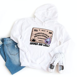 Pu&ograve; includere: Felpa con cappuccio bianca con una grafica di una tavola Ouija con il testo "Summon and Chill?" stampato su di essa.