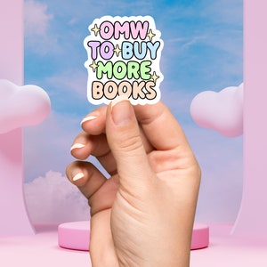 May include: A pastel pink, blue, yellow, and green sticker with the text "OMW TO BUY MORE BOOKS" with stars on either side of the text. The sticker is being held by a hand against a light blue background with pink clouds.
