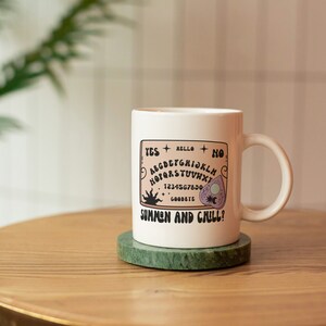 Pu&ograve; includere: Tazza in ceramica bianca con un'illustrazione in bianco e nero di una tavola Ouija con il testo "Summon and Chill?" sul davanti. La tazza &egrave; appoggiata su un sottobicchiere di marmo verde su un tavolo di legno.