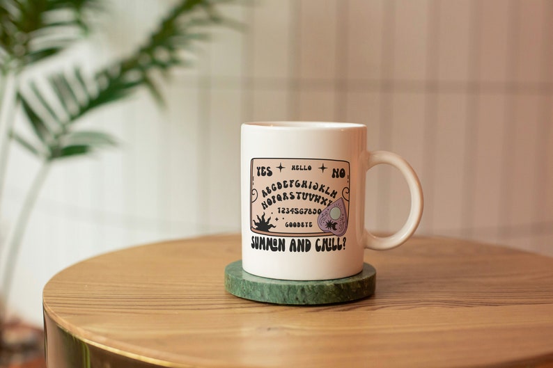 Pu&ograve; includere: Tazza in ceramica bianca con un'illustrazione in bianco e nero di una tavola Ouija con il testo "Summon and Chill?" sul davanti. La tazza &egrave; appoggiata su un sottobicchiere di marmo verde su un tavolo di legno.