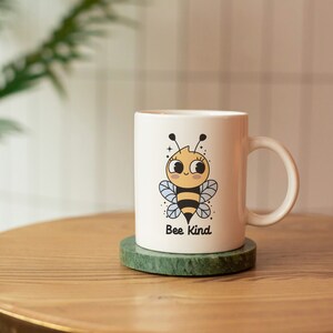 May include: White ceramic mug with a cartoon bee illustration and the text "Bee Kind". The mug is on a green marble coaster on a wooden table.