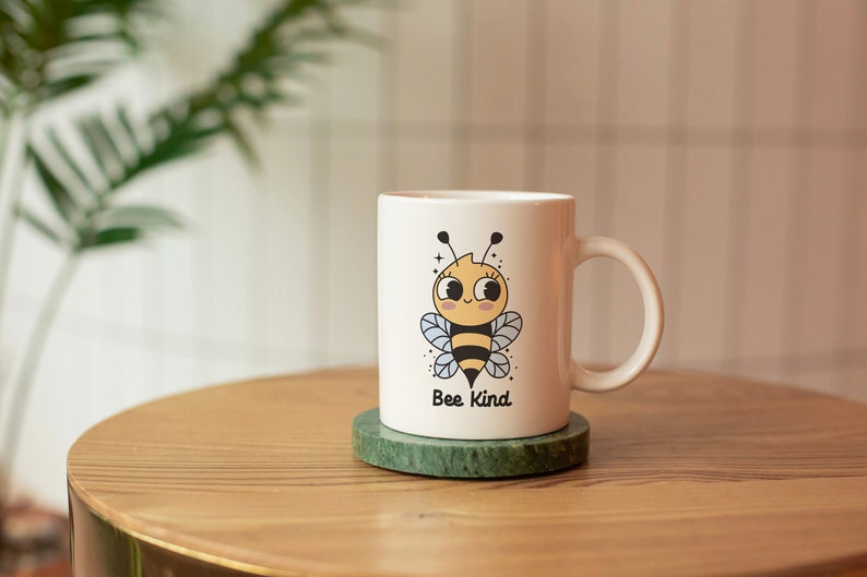 May include: White ceramic mug with a cartoon bee illustration and the text "Bee Kind". The mug is on a green marble coaster on a wooden table.