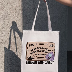 Pu&ograve; includere: Borsa tote bianca con un design di tavola ouija marrone e bianco. La tavola dice "YES", "HELLO", "NO", "GOODBYE", e "SUMMEN AND CHILL?" con una planchette viola.