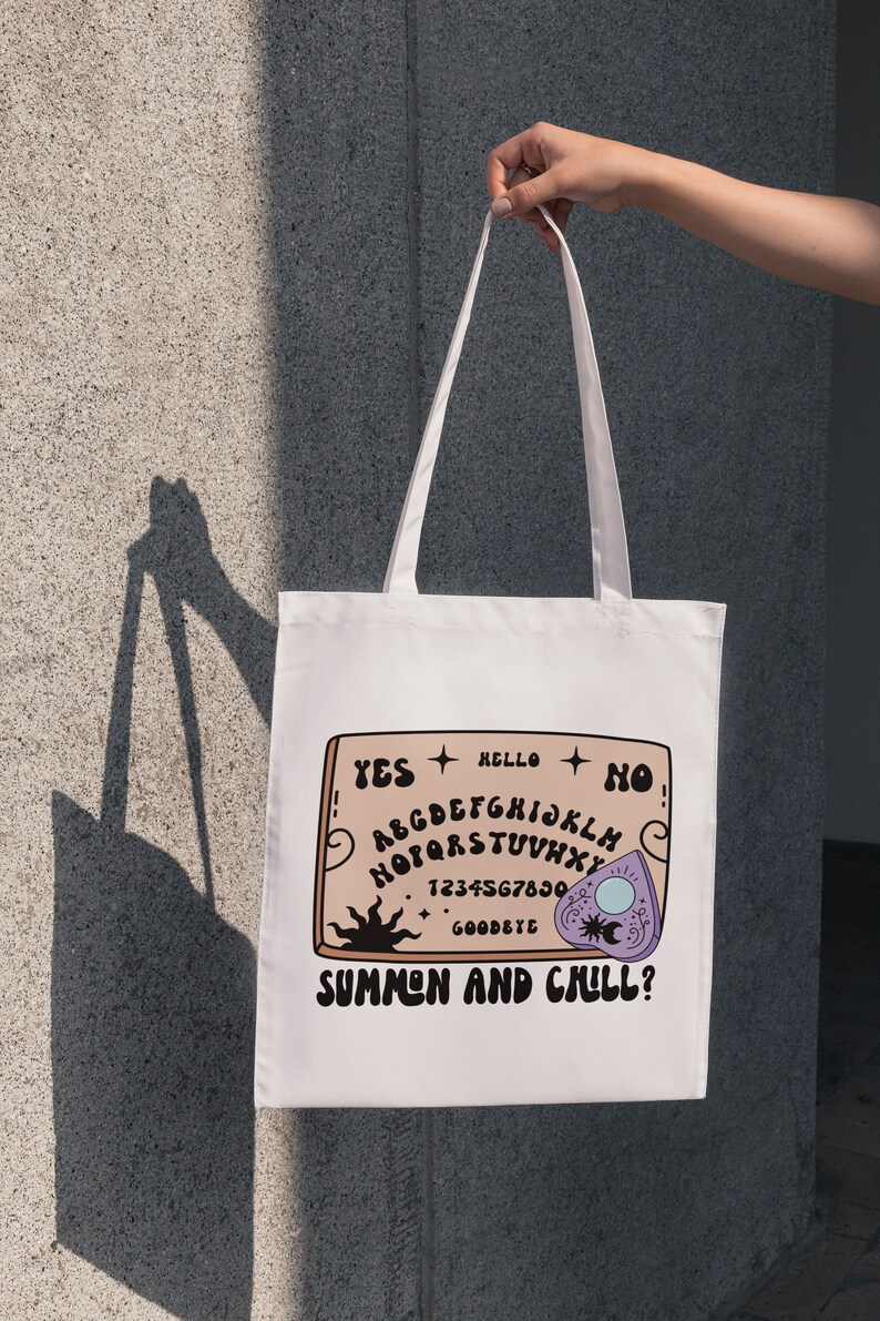 Pu&ograve; includere: Borsa tote bianca con un design di tavola ouija marrone e bianco. La tavola dice "YES", "HELLO", "NO", "GOODBYE", e "SUMMEN AND CHILL?" con una planchette viola.