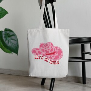 Pode incluir: Uma sacola branca com um chap&eacute;u de cowboy rosa e o texto "LET'S GO GIRLS" em vermelho. A sacola est&aacute; pendurada em uma cadeira preta, com uma planta verde ao fundo.