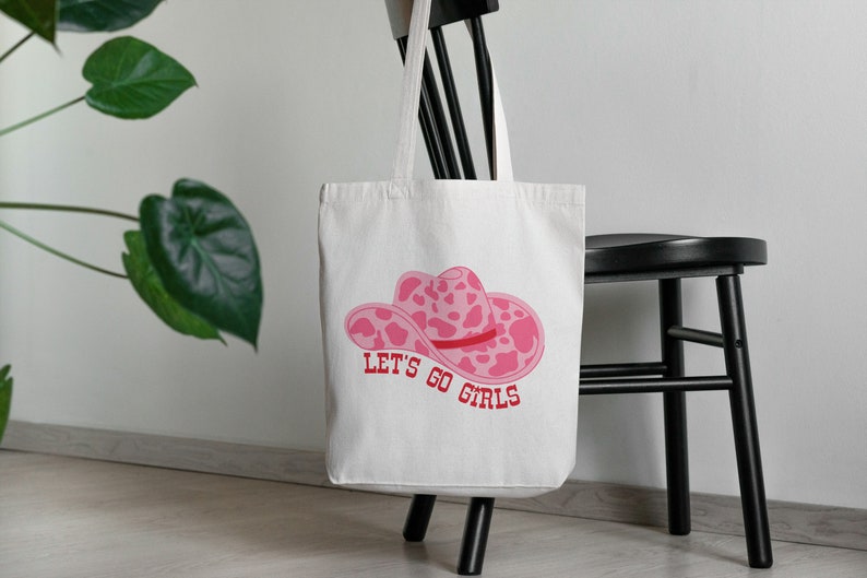 Pode incluir: Uma sacola branca com um chap&eacute;u de cowboy rosa e o texto "LET'S GO GIRLS" em vermelho. A sacola est&aacute; pendurada em uma cadeira preta, com uma planta verde ao fundo.