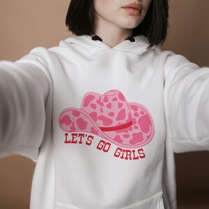 Pode incluir: Moletom branco com um gr&aacute;fico de chap&eacute;u de cowboy rosa com um design de estampa de vaca e o texto "LET'S GO GIRLS" em vermelho. O moletom tem um bolso frontal e um capuz com cord&atilde;o.