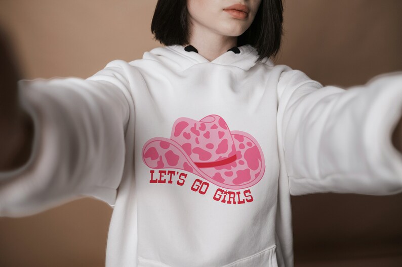 Pode incluir: Moletom branco com um gr&aacute;fico de chap&eacute;u de cowboy rosa com um design de estampa de vaca e o texto "LET'S GO GIRLS" em vermelho. O moletom tem um bolso frontal e um capuz com cord&atilde;o.