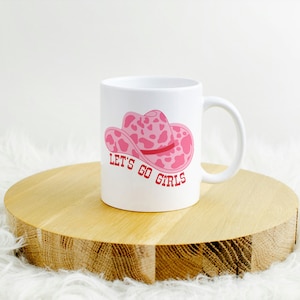 Pode incluir: Caneca de cer&acirc;mica branca com um design de chap&eacute;u de cowboy rosa com estampa de vaca e o texto "LET'S GO GIRLS" em vermelho. A caneca est&aacute; sobre uma superf&iacute;cie de madeira.