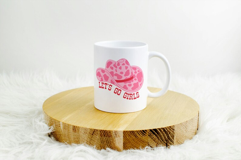 Pode incluir: Caneca de cer&acirc;mica branca com um design de chap&eacute;u de cowboy rosa com estampa de vaca e o texto "LET'S GO GIRLS" em vermelho. A caneca est&aacute; sobre uma superf&iacute;cie de madeira.