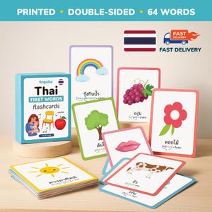 Könnte beinhalten: Ein Set thailändischer Erstwort-Lernkarten mit farbenfrohen Illustrationen. Die Karten zeigen Bilder eines Regenbogens, Trauben, eines Baumes, einer Blume, Lippen, einer Kuh und einer lächelnden Sonne. Die Box trägt die Aufschrift "Thai FIRST WORDS flashcards".
