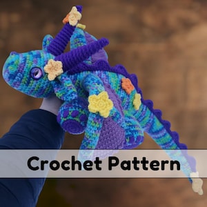 May include: Crochet pattern for a purple and blue dragon with yellow and orange flowers. The dragon is holding a flower in its mouth.