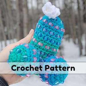 May include: A crochet pattern for a blue and pink crocheted toy with a white flower on top. The toy is shaped like a heart with two rounded ends.