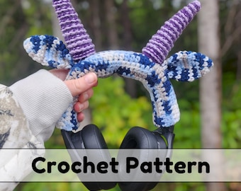 May include: A handmade crochet headphone accessory with a horn and wing design. The accessory is primarily blue, gray, and white, with purple horns. The text "Crochet Pattern" is displayed on the image. The accessory is designed to fit over a pair of black headphones.