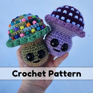 May include: Two crocheted mushroom amigurumi toys. One is green with colourful beads on the top and the other is purple with light blue beads. Both have black eyes and a smiling mouth.