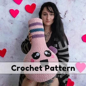 May include: A pink crochet plushie with a cartoon face and striped neck. The plushie has black eyes, a black mouth, and pink cheeks. The text "Crochet Pattern" is displayed on the bottom of the image. Hearts are in the background.