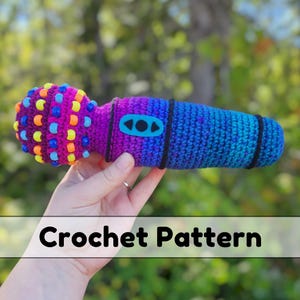 May include: A handmade crochet microphone in shades of purple and blue, adorned with colorful beads. The microphone has a black band and a blue button. The text "Crochet Pattern" is displayed at the bottom of the image.